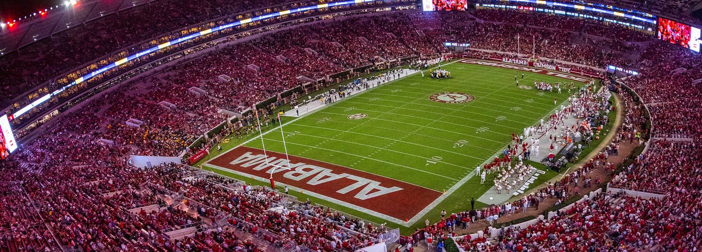 Wide aerial view of Bryant-Denny Stadium at night, the stands filled with crimson and the Alabama end zone lettering visible below