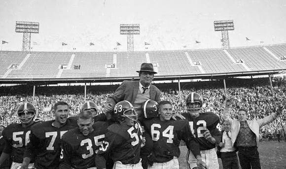 Black and white photograph of Alabama players in leather-era helmets carrying their coach from the field in front of a packed stand