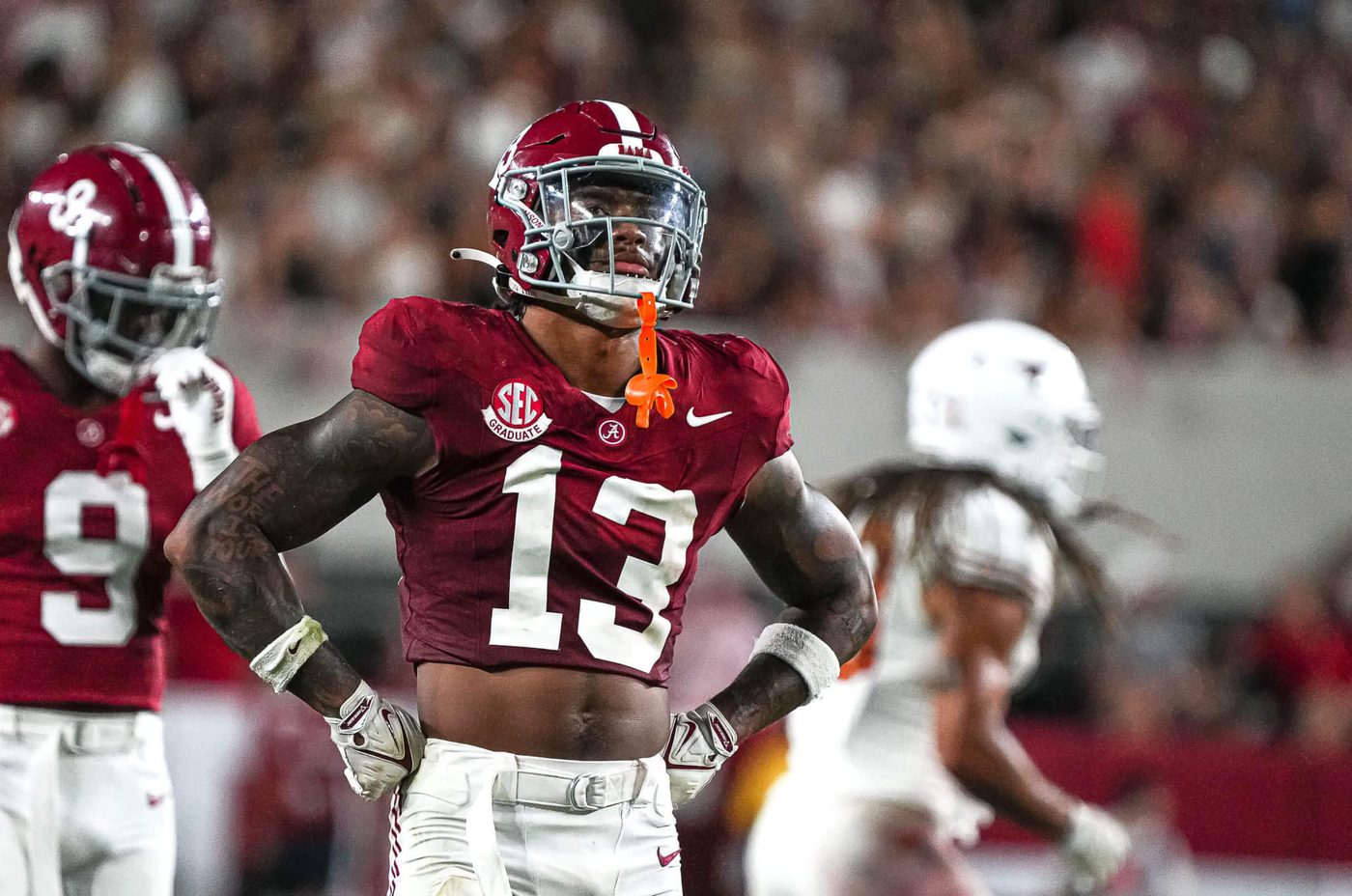 An Alabama defensive back in a crimson jersey standing with hands on hips between plays as a teammate looks on