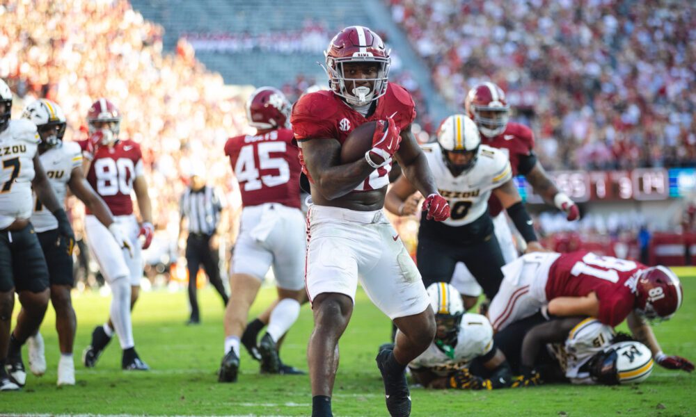 An Alabama running back in crimson carrying the ball into the end zone past Missouri defenders in white and gold