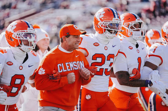 A Clemson Tigers football coach standing on the sideline with players wearing orange jerseys and white numbers, speaking to the team during a game. The coach demonstrates leadership and strategy during live game action.