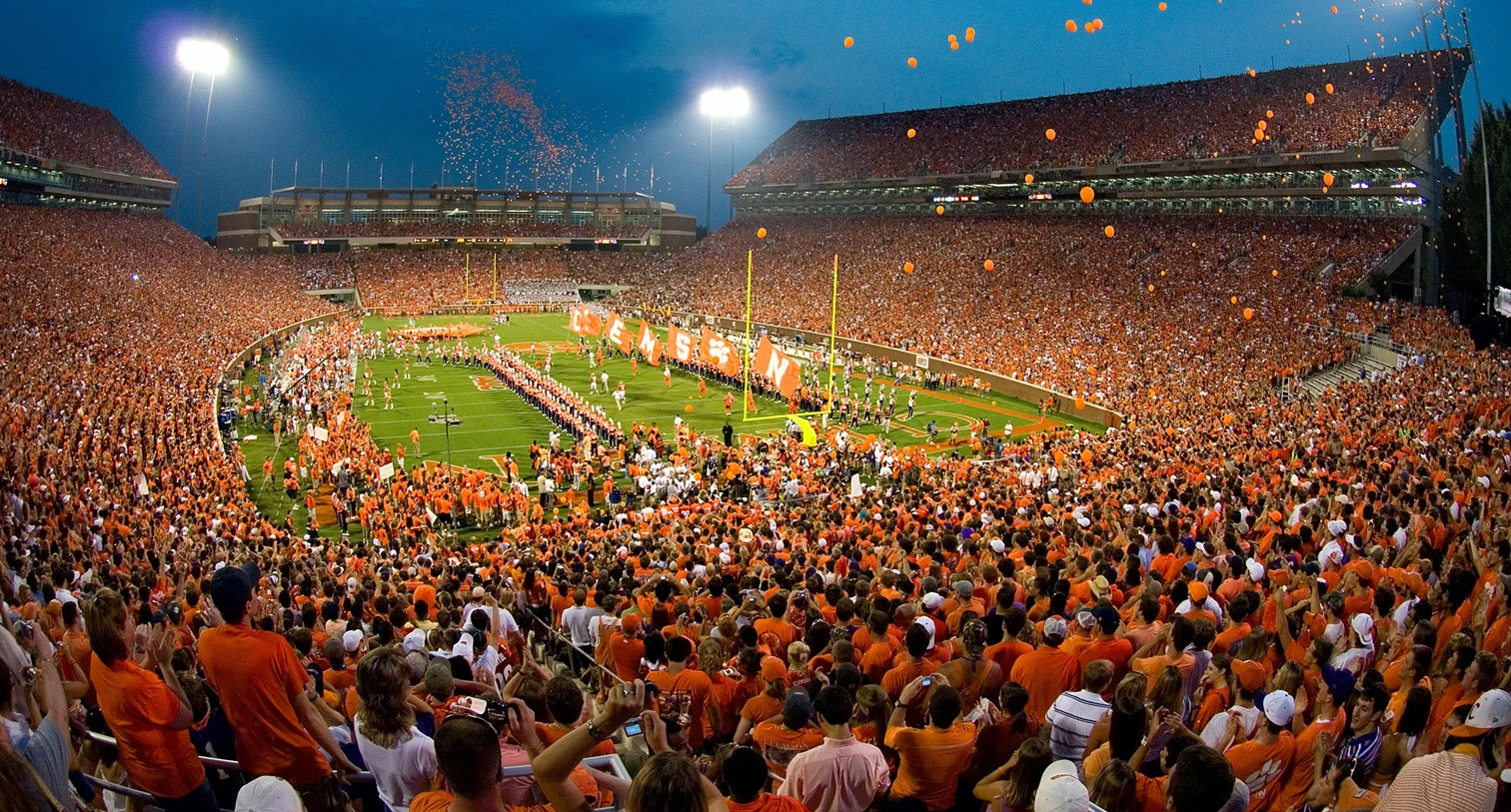 Memorial Stadium in Clemson, South Carolina, known as Death Valley, filled with thousands of passionate Clemson Tigers supporters wearing orange under the stadium lights during a night game. The venue is packed with fans and confetti falls from the stands creating an electric atmosphere.