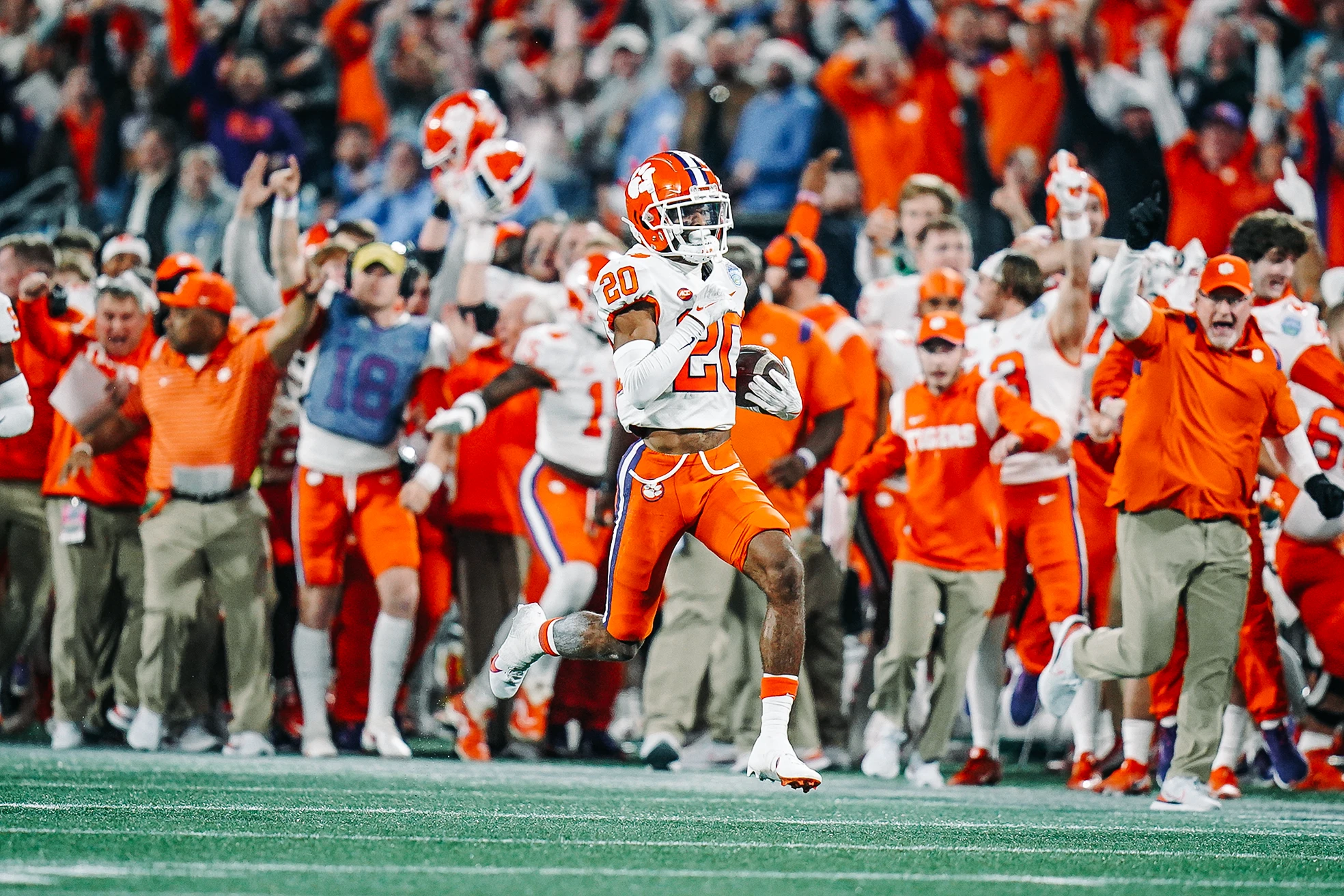 A Clemson Tigers player wearing an orange jersey running powerfully with the football down the field, evading defenders as fans in the background cheer and celebrate the exciting run.