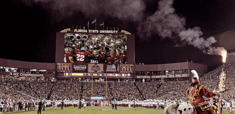 Doak Campbell Stadium in Tallahassee, Florida on game night, filled with thousands of Florida State Seminoles supporters. The field features the iconic flaming spear tradition with thousands of fans in garnet and gold creating an intimidating home-field atmosphere.