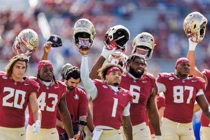 Florida State Seminoles football players standing together holding their garnet and gold helmets raised up in the air, displaying team unity, brotherhood, and celebration after a successful play.
