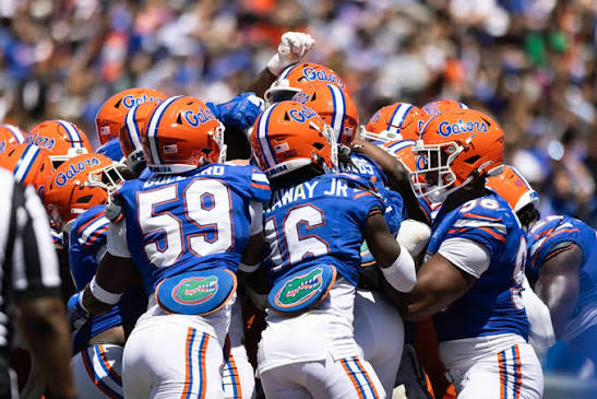 Florida Gators football players standing close together in a huddle formation wearing blue jerseys and helmets with the Gators logo, gathered to hear the play call and prepare for the next snap.