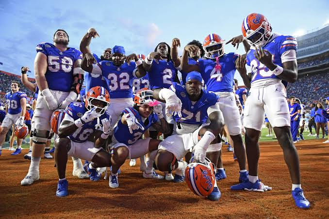 Florida Gators football players wearing blue jerseys gathered closely together on the field celebrating a successful play or score, with fans visible cheering in the background. The players demonstrate camaraderie and excitement during game action.