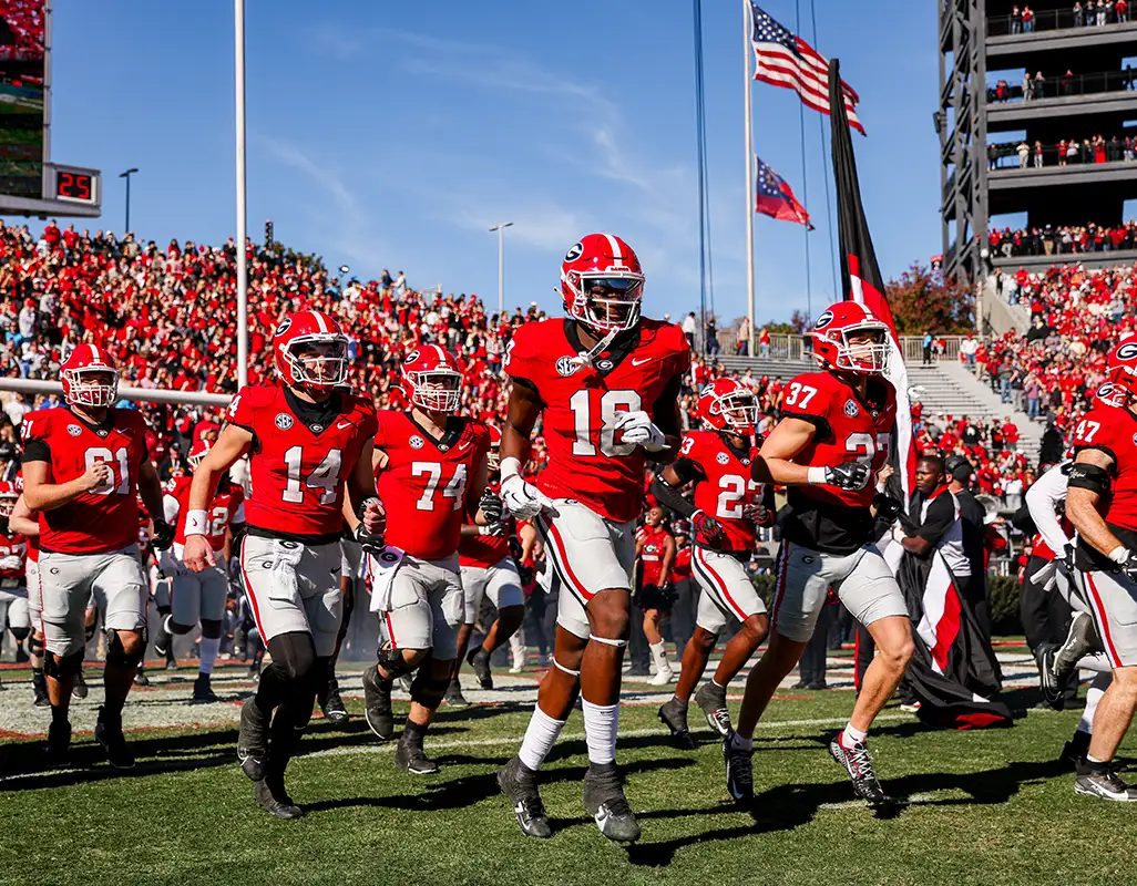 Georgia players jogging onto the field on a clear autumn day, American and state flags flying behind the stand