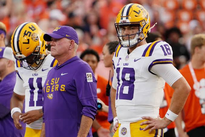 An LSU quarterback in a white number 18 jersey standing with hands on hips beside the head coach in a purple LSU Tigers pullover before a road game