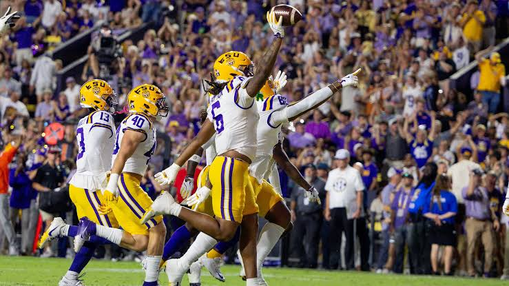 An LSU defender holding the football aloft in one hand as teammates in white and gold celebrate a turnover in front of a full night crowd