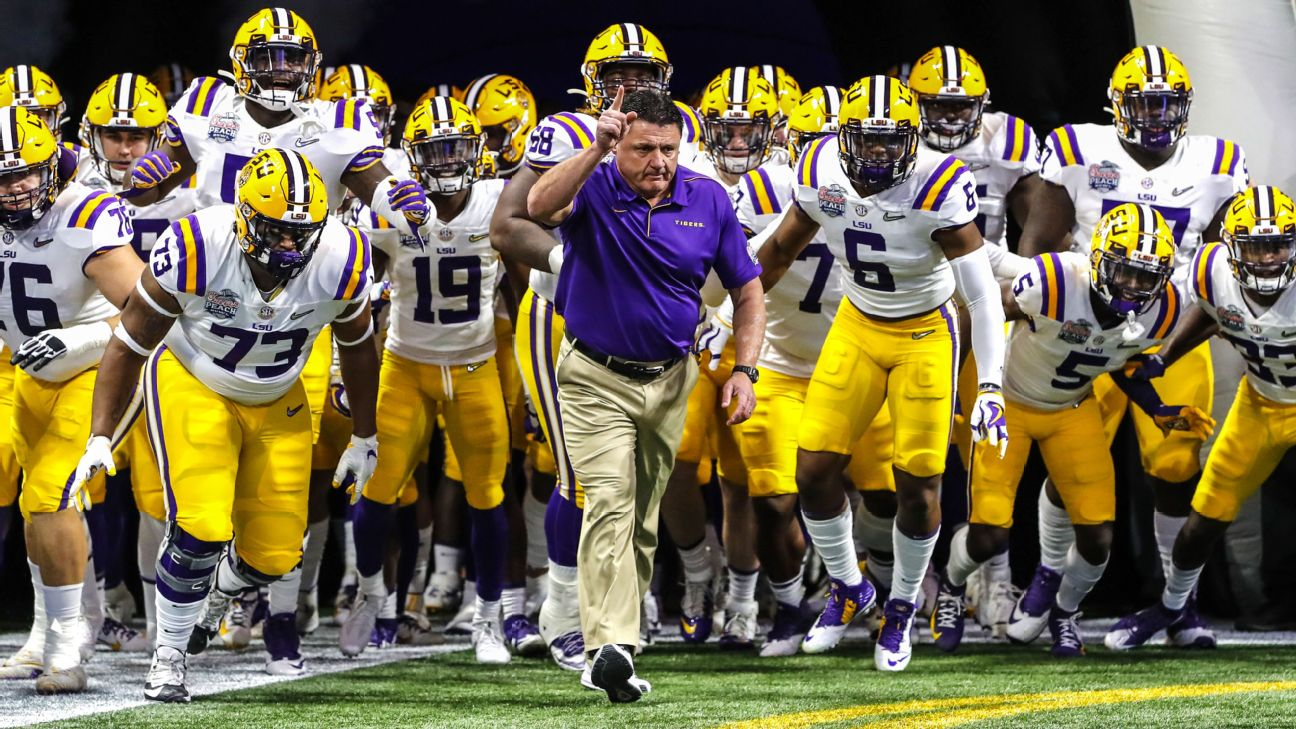 LSU players in white jerseys and gold helmets running out of the tunnel behind their head coach, who holds one finger raised