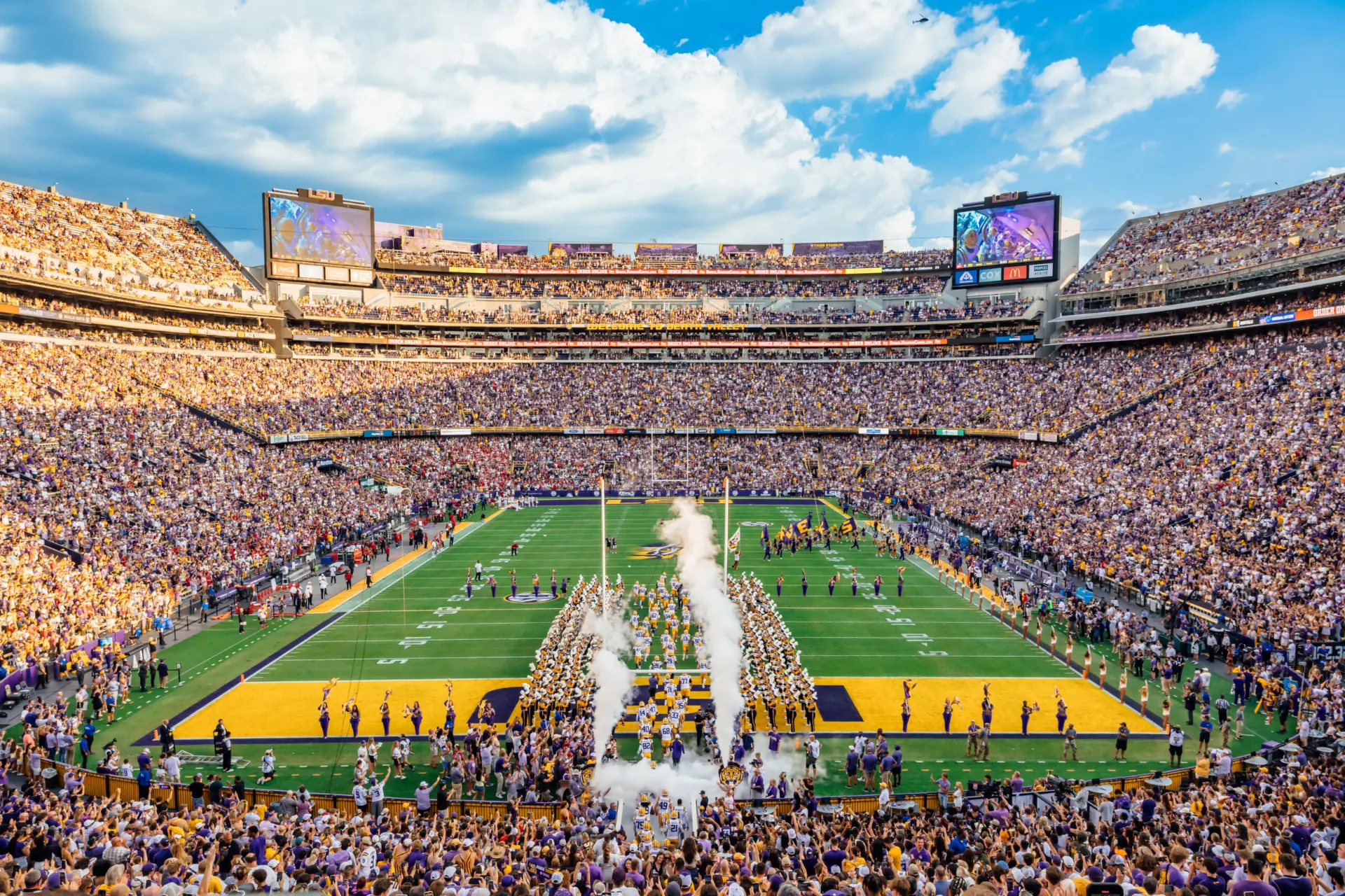Tiger Stadium packed with supporters in purple and gold as LSU players run onto the field through smoke, the band formed up in a tunnel on the grass
