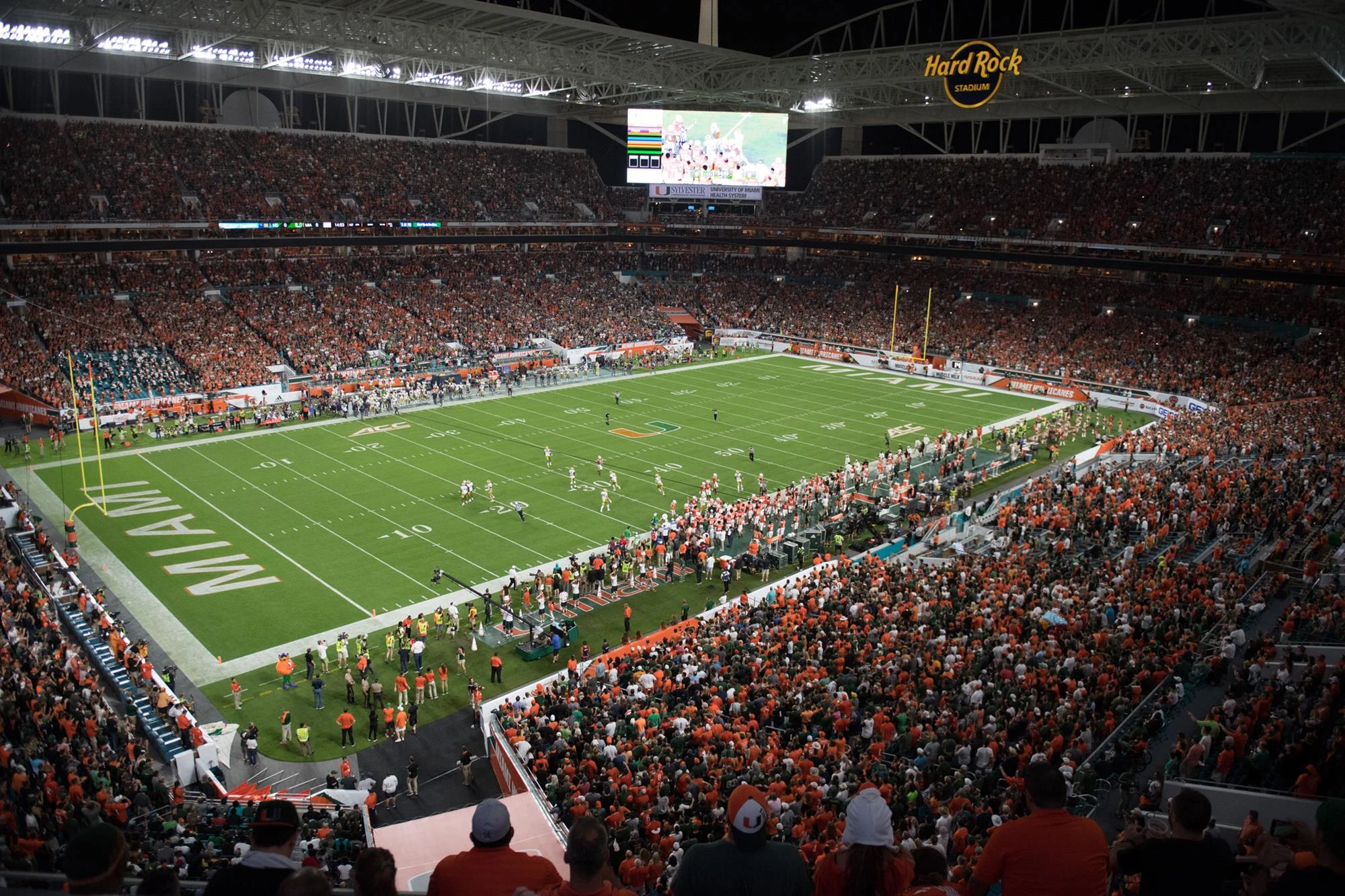 Hard Rock Stadium in Miami, Florida on game night filled with thousands of Miami Hurricanes supporters under the stadium lights. The venue is packed with fans wearing orange and green creating an energetic atmosphere for Hurricanes football.