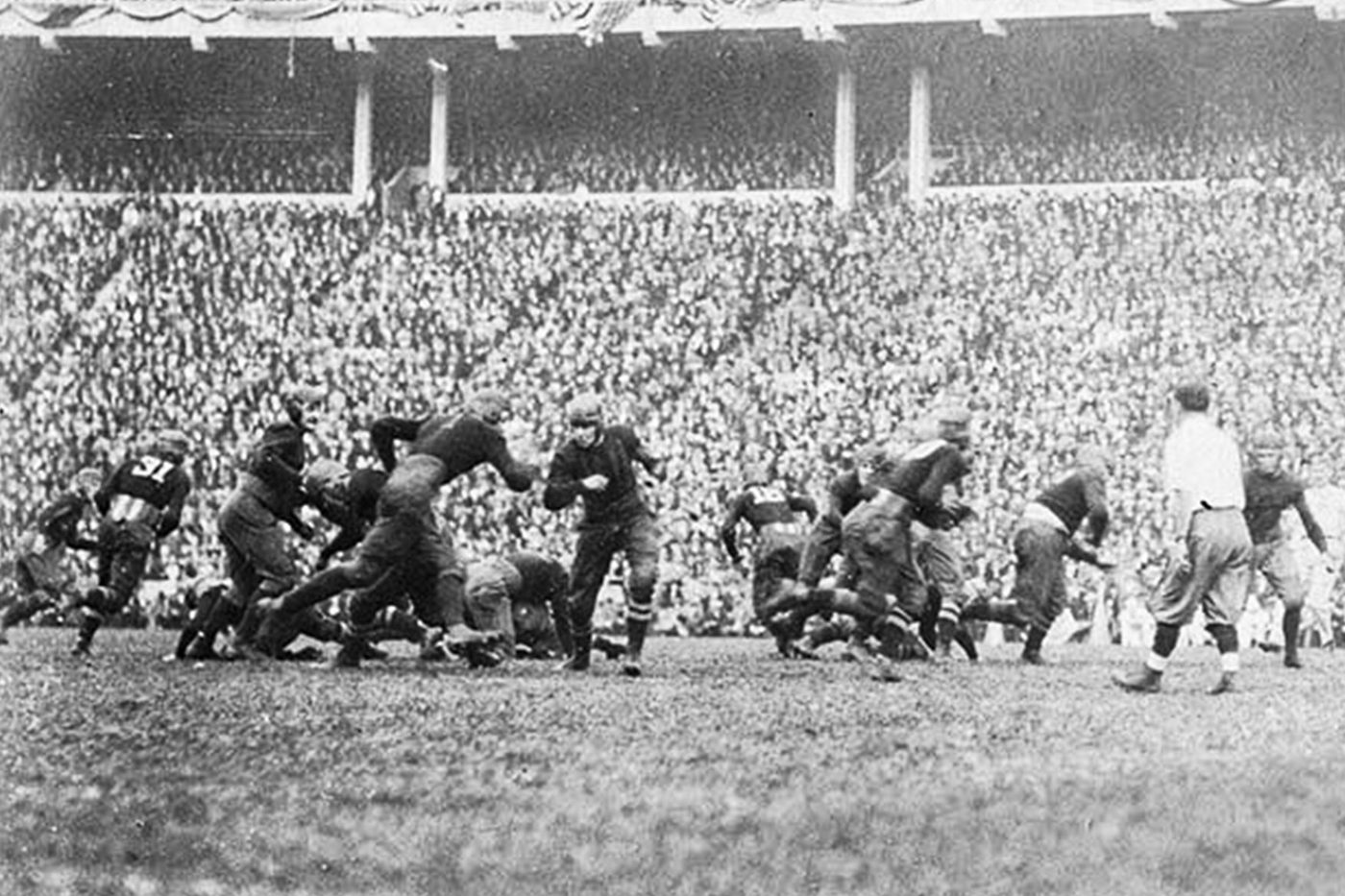 Black and white photograph of early era college football, players in leather helmets contesting a run in front of a vast packed grandstand