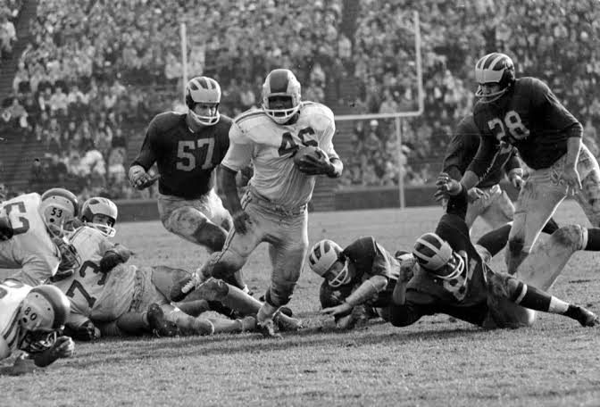 Black and white mid-century game action, a ball carrier in a light jersey breaking through the line past defenders in dark jerseys