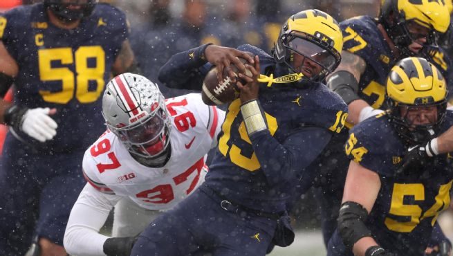 A Michigan quarterback in navy being wrapped up by an Ohio State defensive lineman in the rain