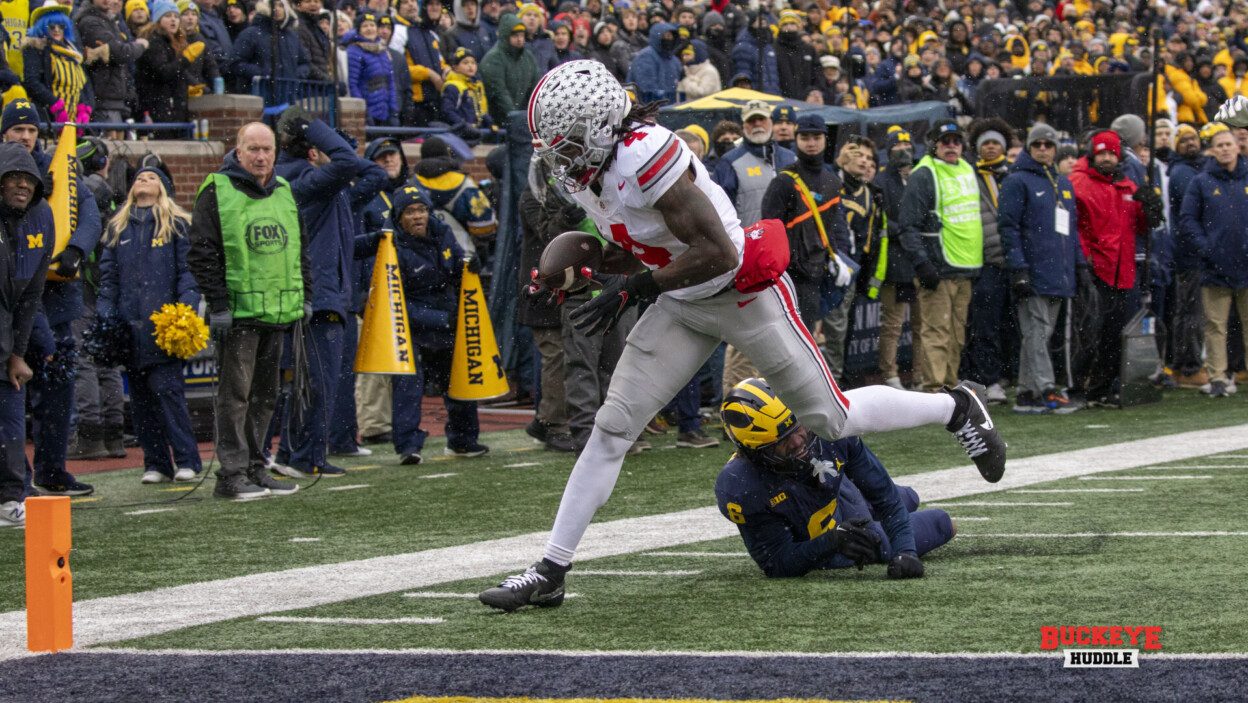 An Ohio State receiver in white catching a touchdown at the pylon in falling snow while a Michigan defender lies on the turf behind him