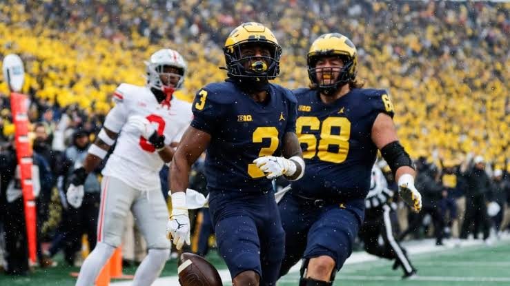 A Michigan running back in navy and gold crossing the goal line with a teammate alongside him as an Ohio State defender trails