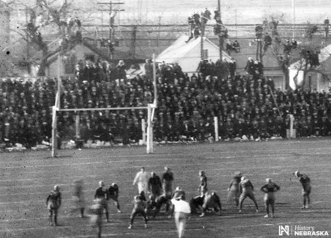 Black and white photograph of early Nebraska football game action, with spectators standing along the sideline and watching from trees behind the goalposts