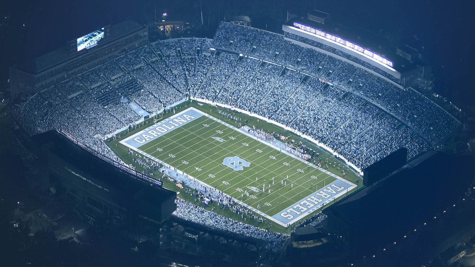 An aerial view of Kenan Stadium in Chapel Hill, North Carolina showing the entire football field and stands packed with thousands of Tar Heels supporters under the lights, with surrounding campus buildings visible.