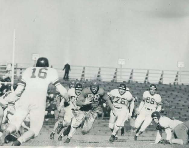 Black and white mid-century game action, a ball carrier in a dark jersey running through tacklers in front of open stadium seating