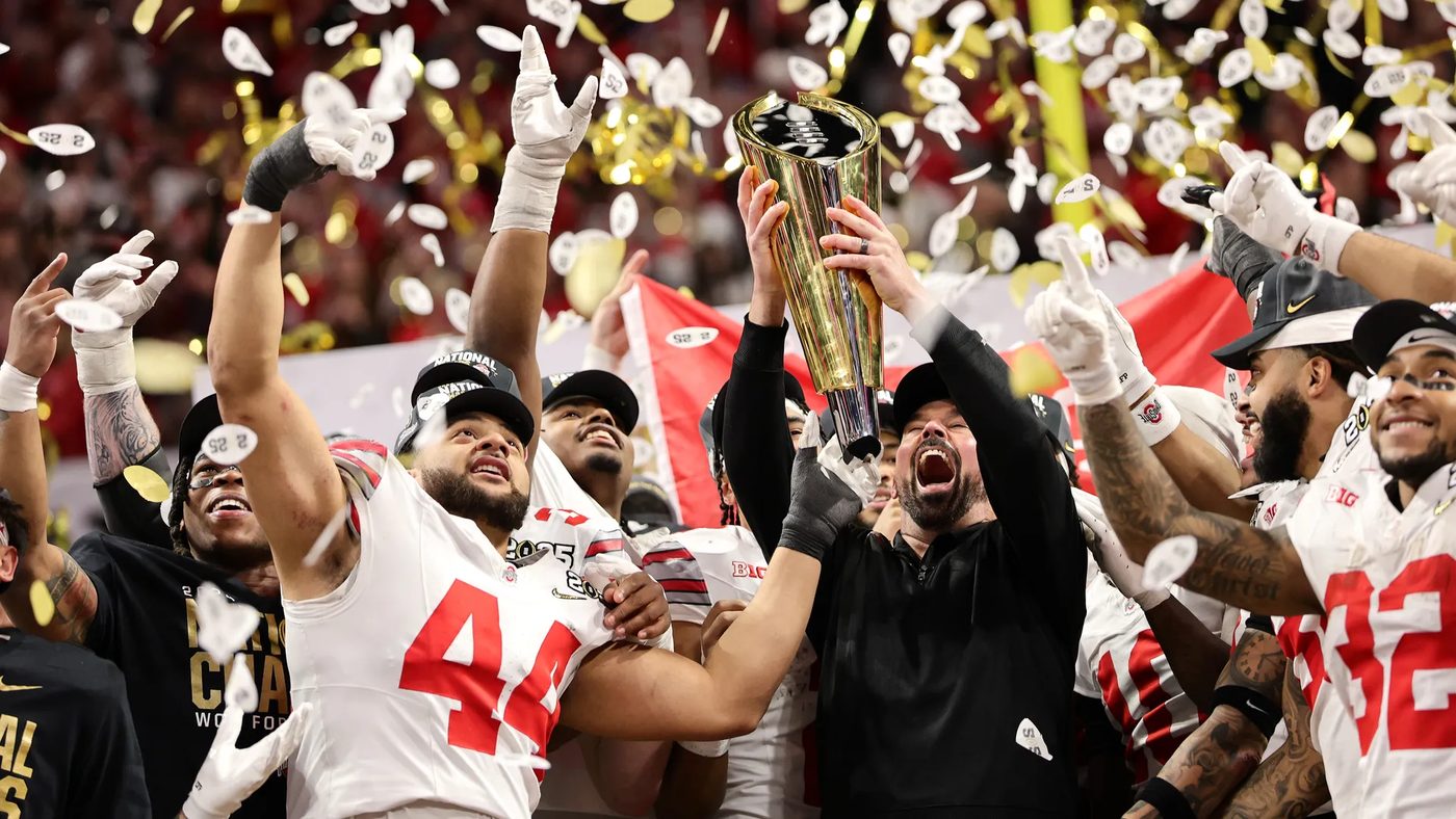 Ohio State players and coaches in white uniforms raising the College Football Playoff national championship trophy amid falling confetti