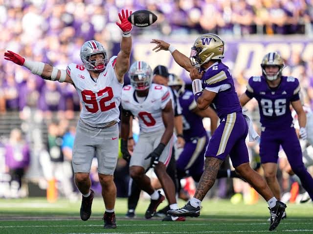 An Ohio State defensive lineman in grey and red raising both arms to bat down a pass from a Washington quarterback in purple