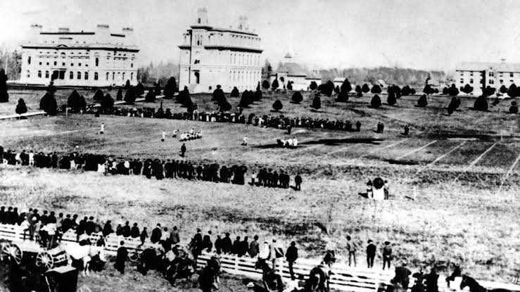 Distant black and white photograph of an early football game on an open campus field, spectators standing in long lines along both touchlines with university buildings behind