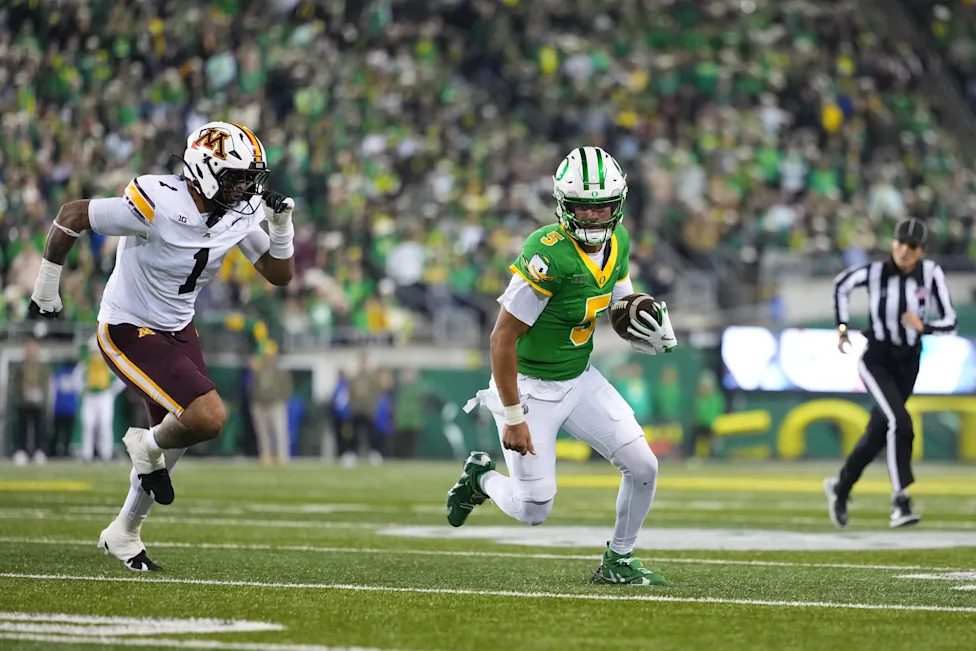 An Oregon quarterback in a green jersey running with the ball down the sideline as a Minnesota defender in white pursues