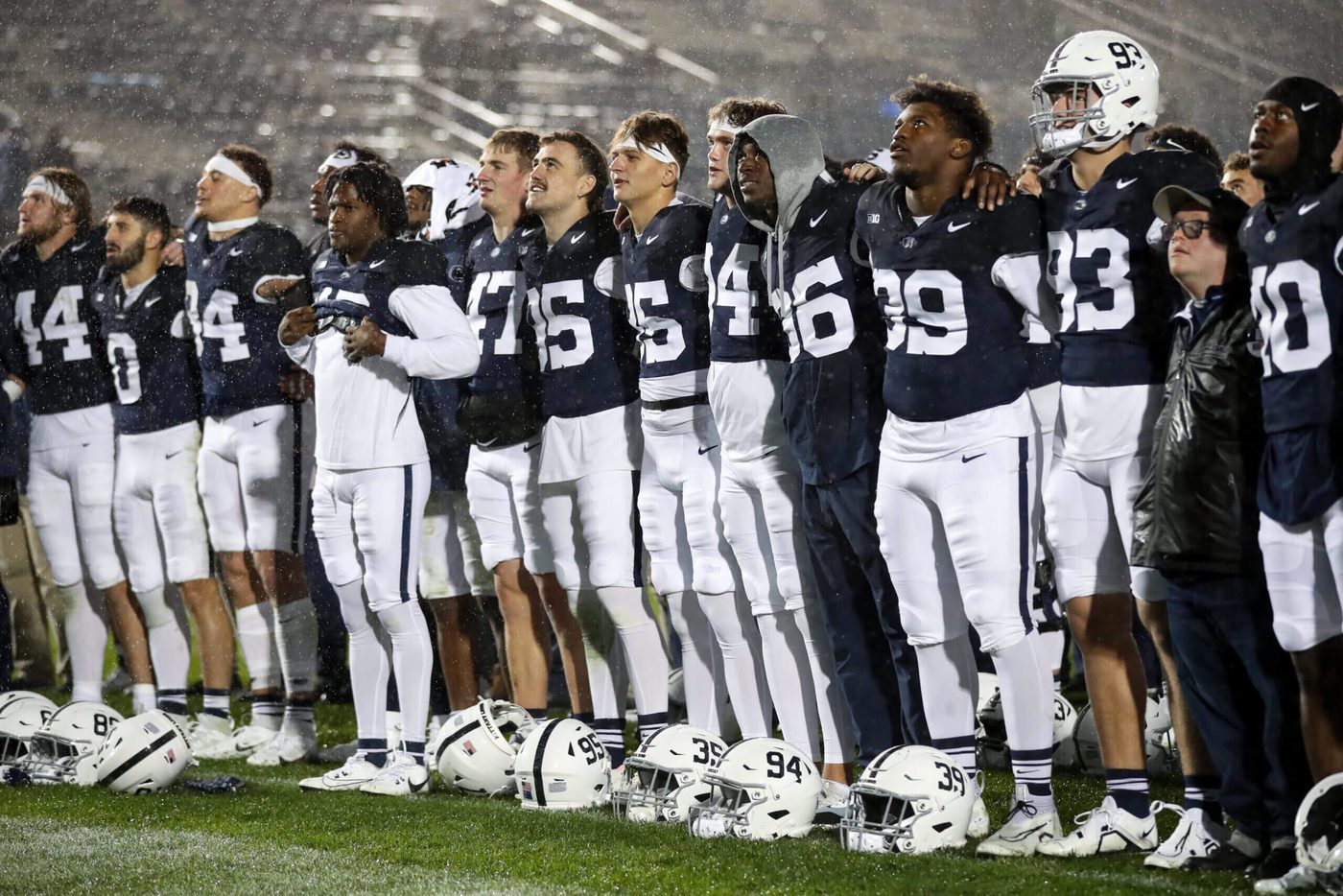 Penn State players standing arm in arm in heavy rain under floodlights, helmets lined up on the grass in front of them