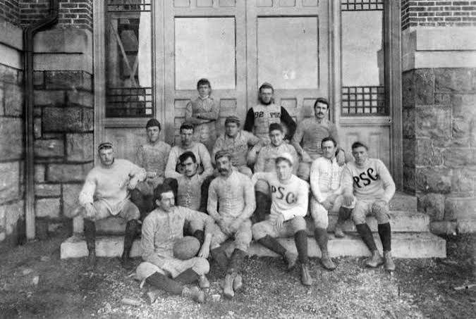 Black and white photograph of an early Penn State team seated on stone steps outside a campus building, one player holding a football