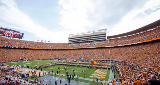 Neyland Stadium in Knoxville, Tennessee on game day filled with tens of thousands of Tennessee Volunteers supporters wearing orange and white, with the Tennessee River visible in the background. The iconic football venue is packed with fans creating a sea of orange throughout the stands during a home game.