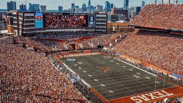 Aerial view of a packed Darrell K Royal-Texas Memorial Stadium in burnt orange, with the Austin skyline rising behind the north end