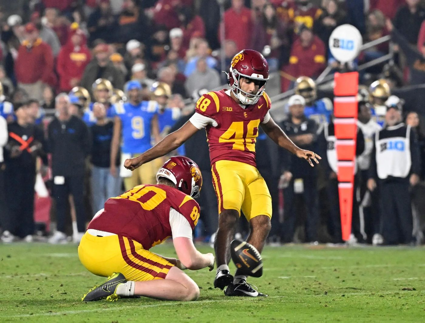A USC kicker striking a field goal from a holder as UCLA players in blue watch from the sideline behind him
