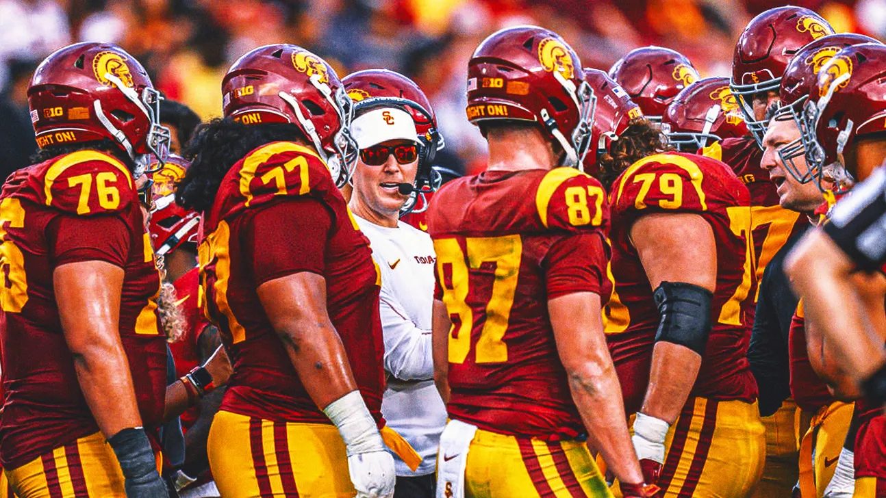 USC head coach in a white cap and shirt speaking to players wearing cardinal jerseys and gold helmets in a sideline huddle